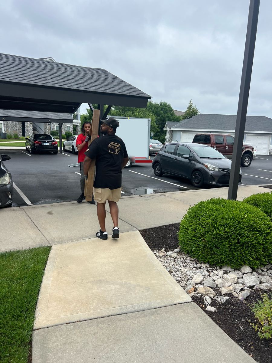 Two crew members carrying a long wooden piece across an apartment parking lot