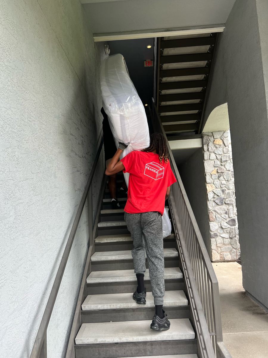 Crew member in a red FRAGILE shirt carrying a wrapped mattress up an outdoor apartment staircase