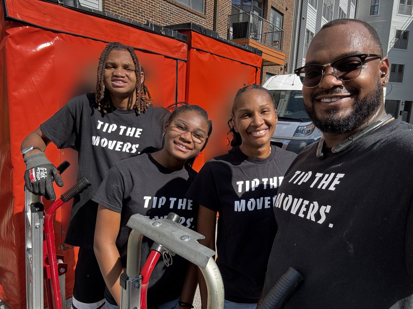 Four Fragile Service Care crew members smiling in black TIP THE MOVERS. shirts with hand trucks on a moving day